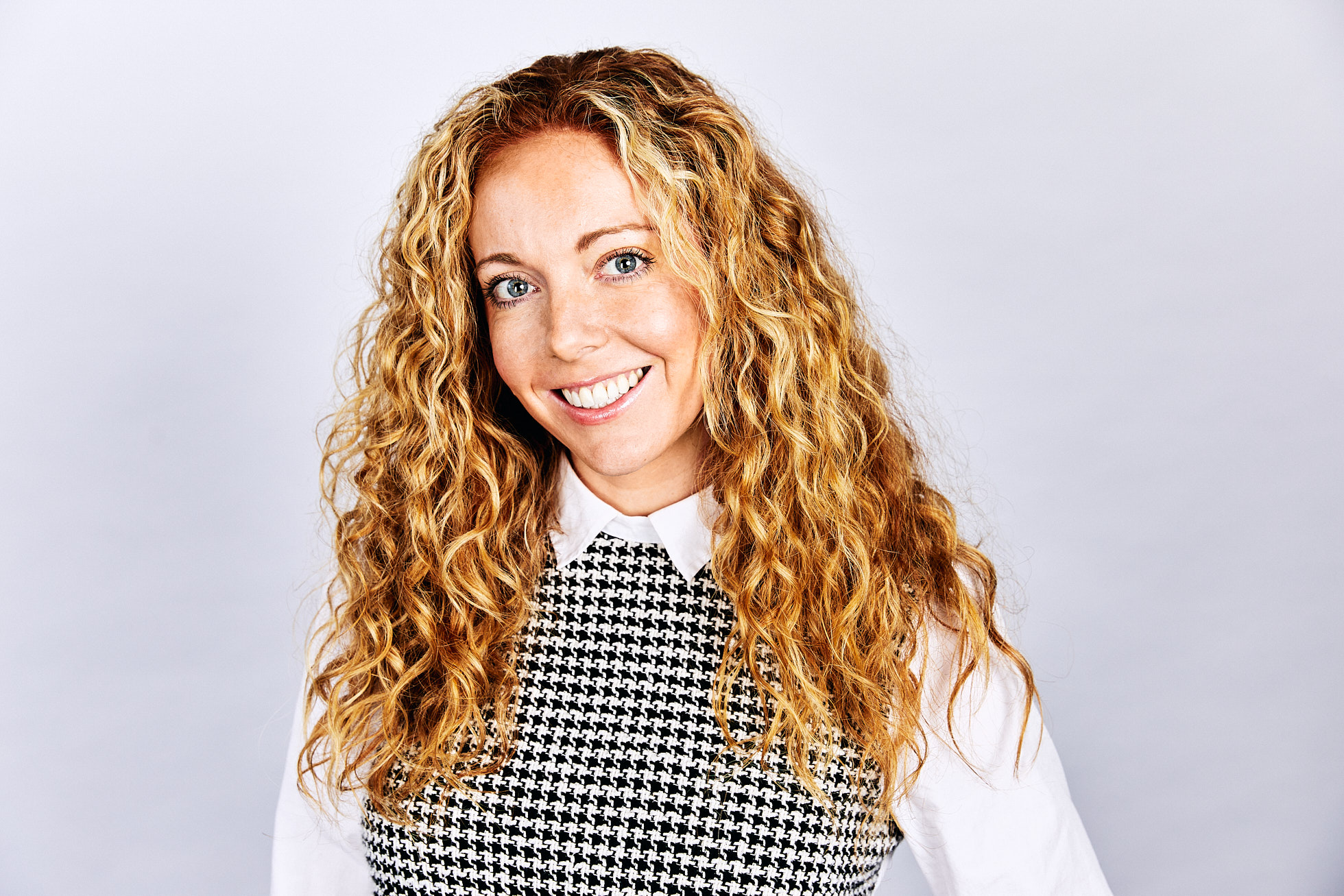 Business headshot of young professional woman with long dark blonde curly hair.  Wearing white blouse with collar and black and white checked dress