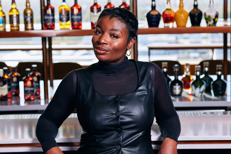Location headshot of woman in black polo with leather dress posing casually in front of a bar