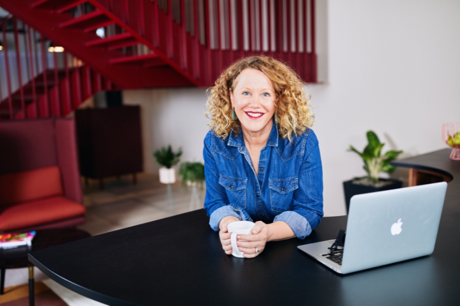 Photo of a woman in a modern business setting with red staircare in the background, she is wearing a denim shirt and leaning on a desk with a laptop open