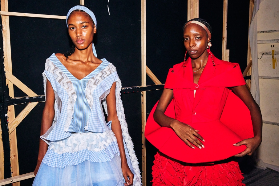 Two models backstage at a catwalk show.  One in light blue long dress and one in red dress