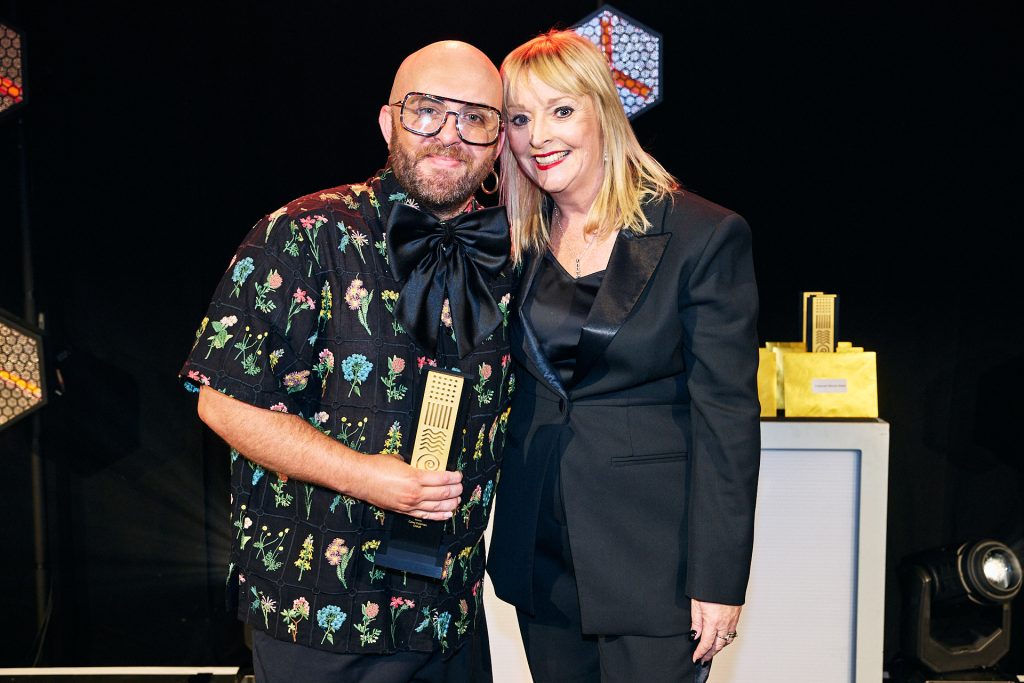 Male with glasses and beard accepting an award from a woman dressed in black