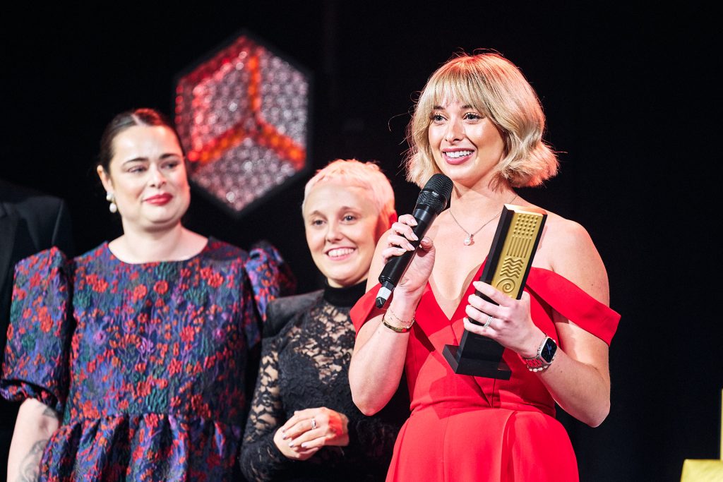 Young woman in red dress accepting an award with two people behind her