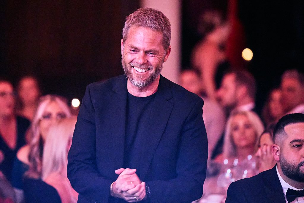 Man in black tee shirt and black suit smiling with this hands clasped walking to accept an award