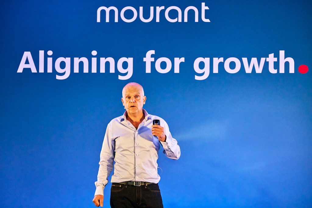 Male speaker in light blue shirt at conference talking on stage with branded blue background