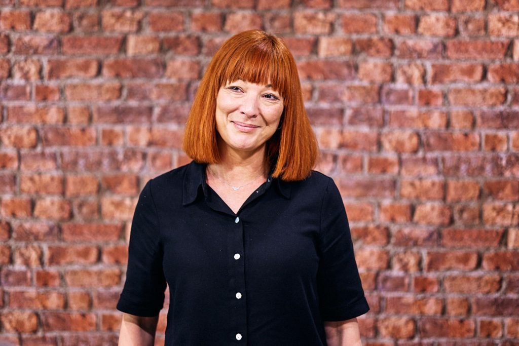 Headshot of professional woman with red hair in a bob, wearing a black top, looking slightly off camera. Shot against a brick background