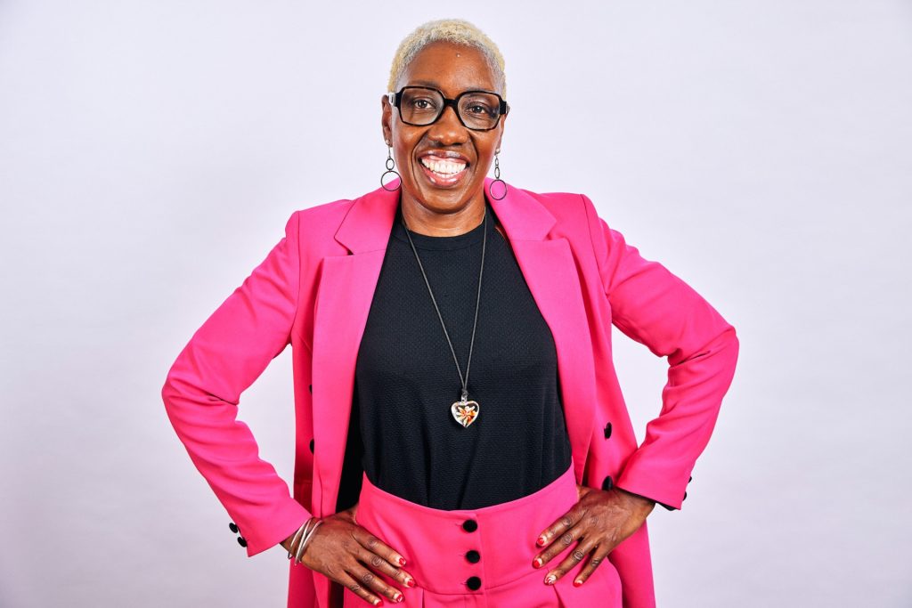 Headshot of a woman with bold black framed glasses, with hands on hips wearing a bright pink suit and black t shirt, smiling for the camera.