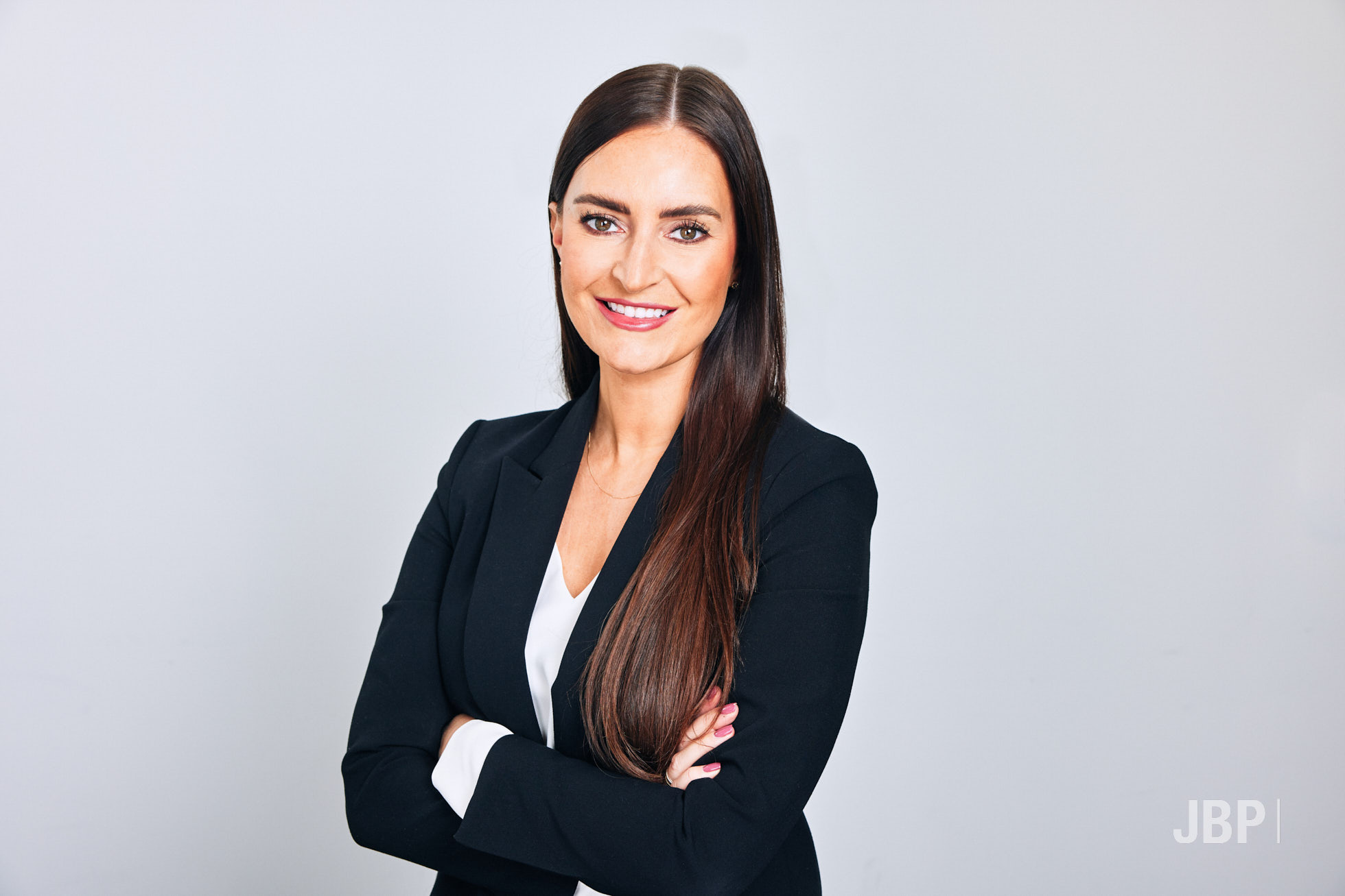Professional Business Headshots by Jon Bradley Photography Business headshot of a young professional woman wearing black blazer and white top with arms folded against a light grey background.