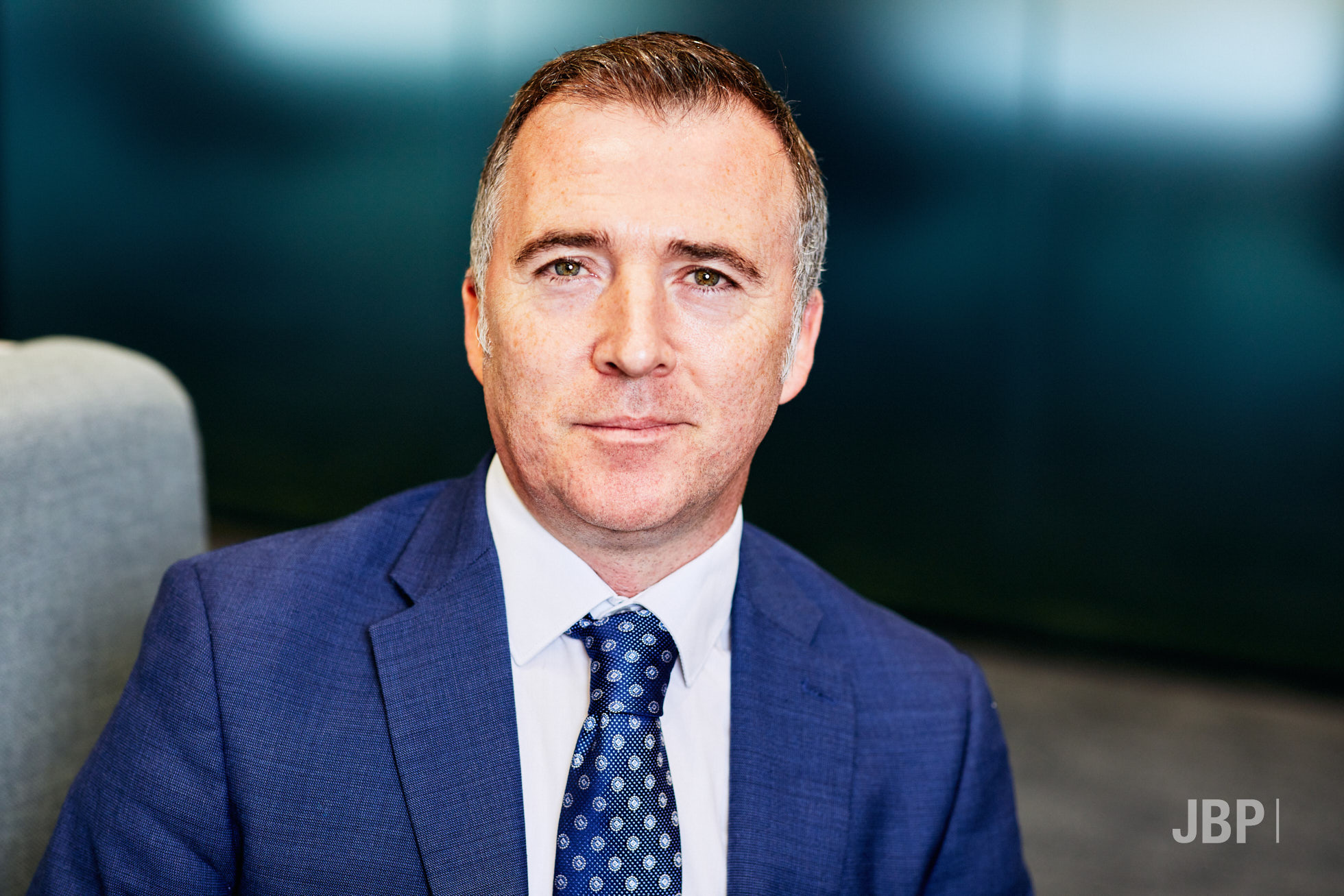 Professional headshot of a man wearing a blue jacket and tie, and a white shirt, against a dark background.
