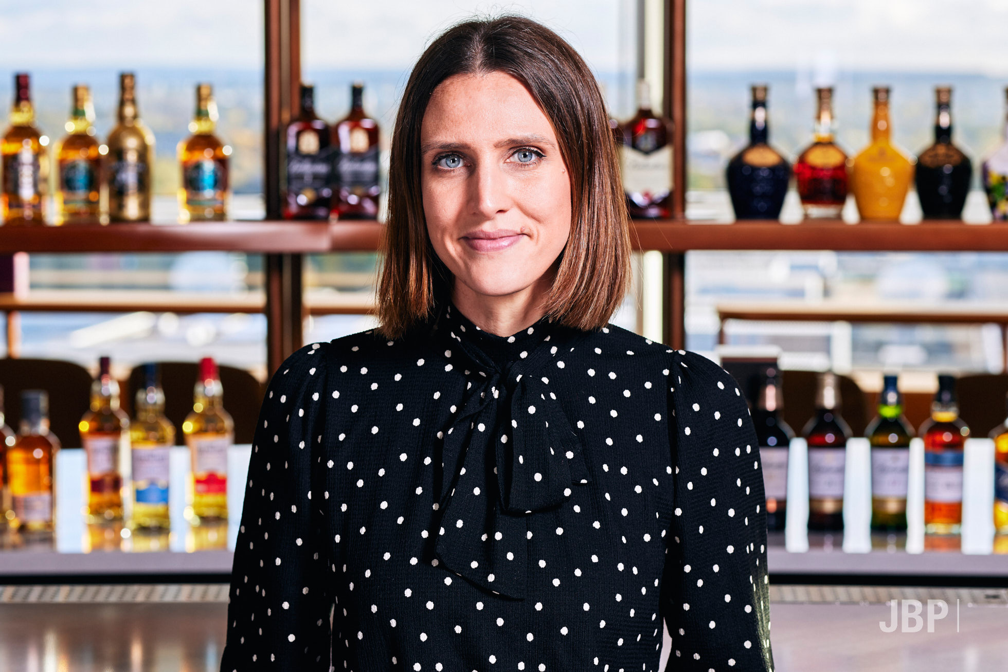 Professional headshot of a woman with brown shoulder length hair wearing a black and white top against a bar backdrop.