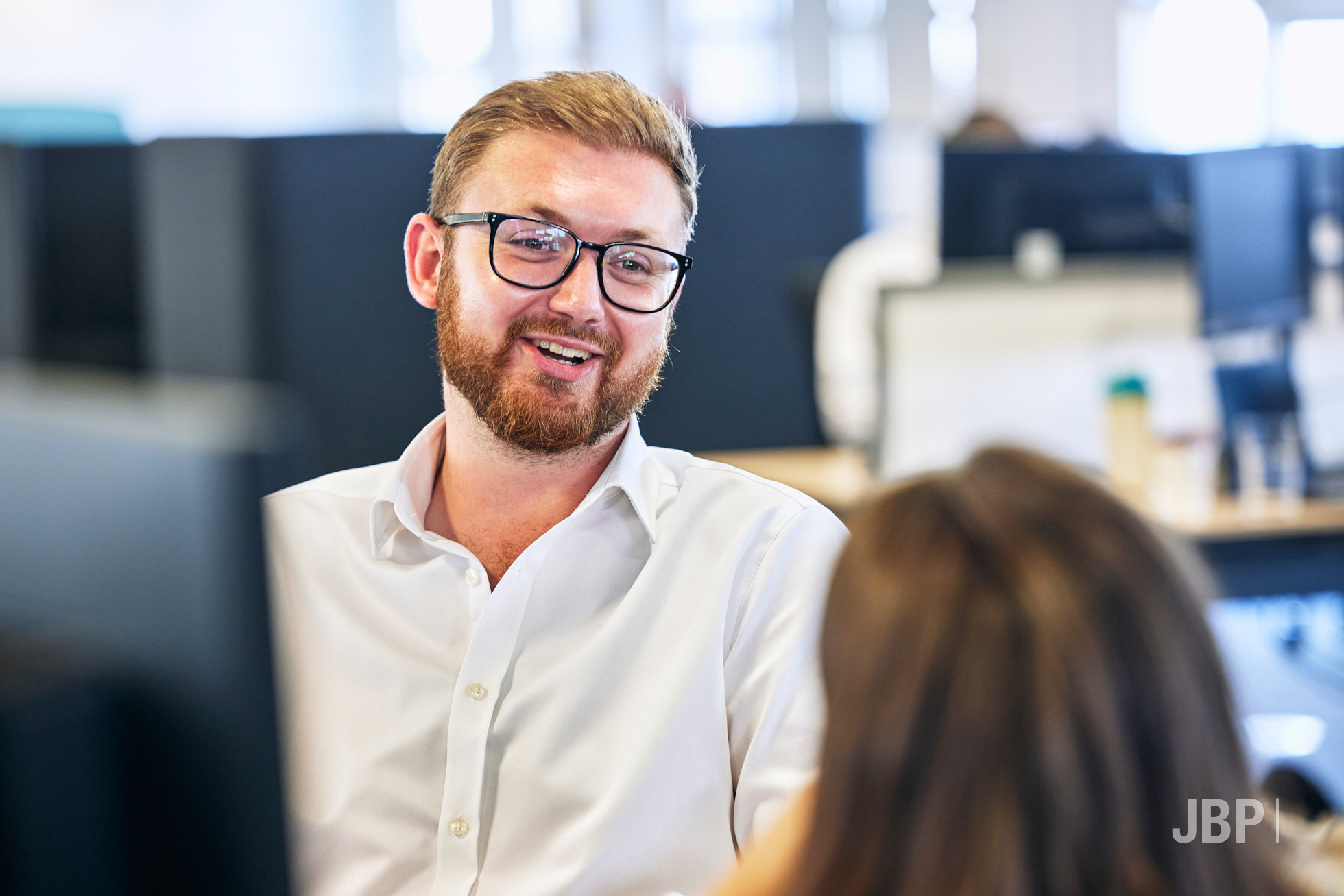 Corporate lifestyle photography showing a smiling with his computer in the corner foreground. He's chatting to a female colleague and the office is out of focus in the background.