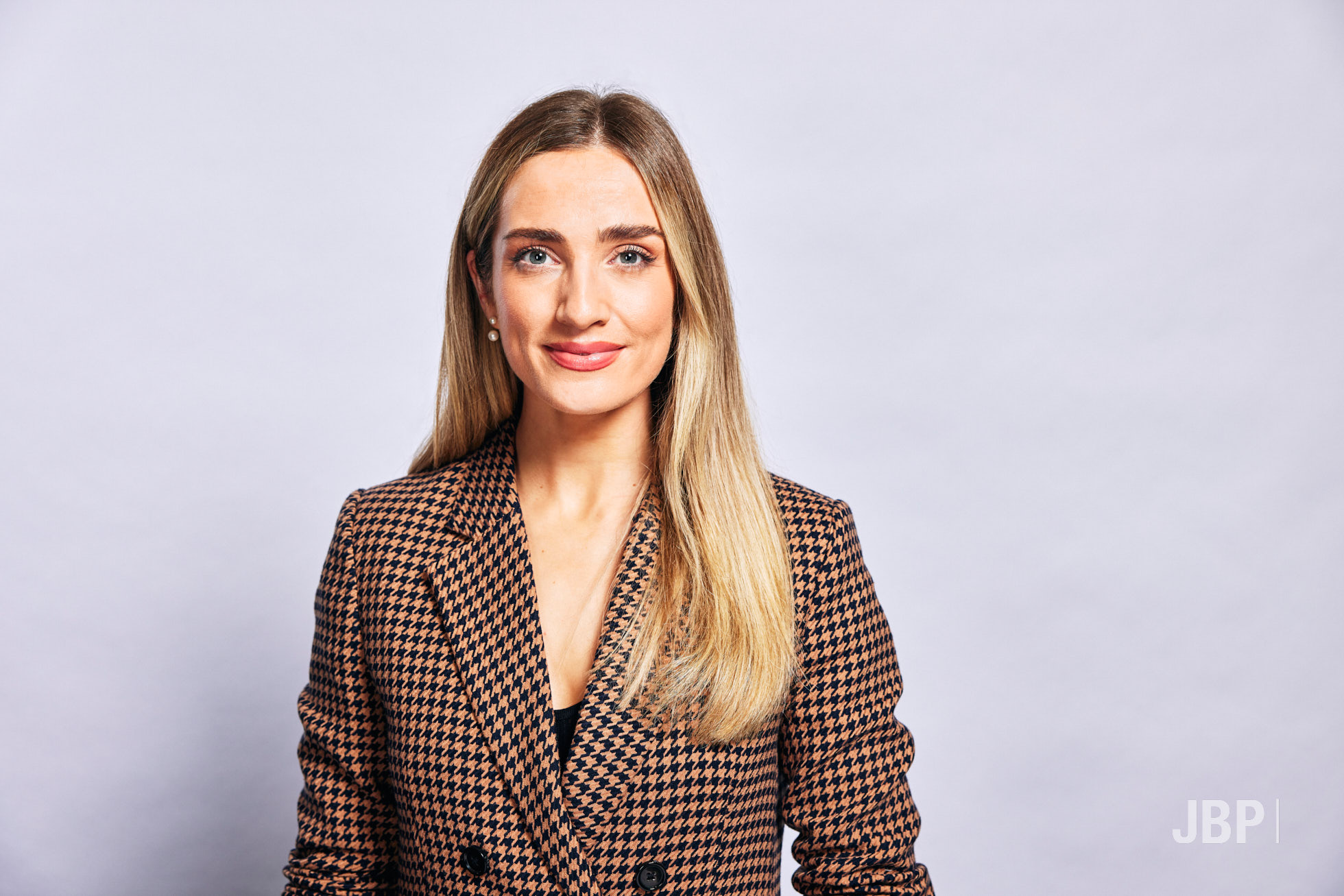 Professional portrait of a woman with long blonde hair dressed in brown adn black checked blazer against a light grey background.