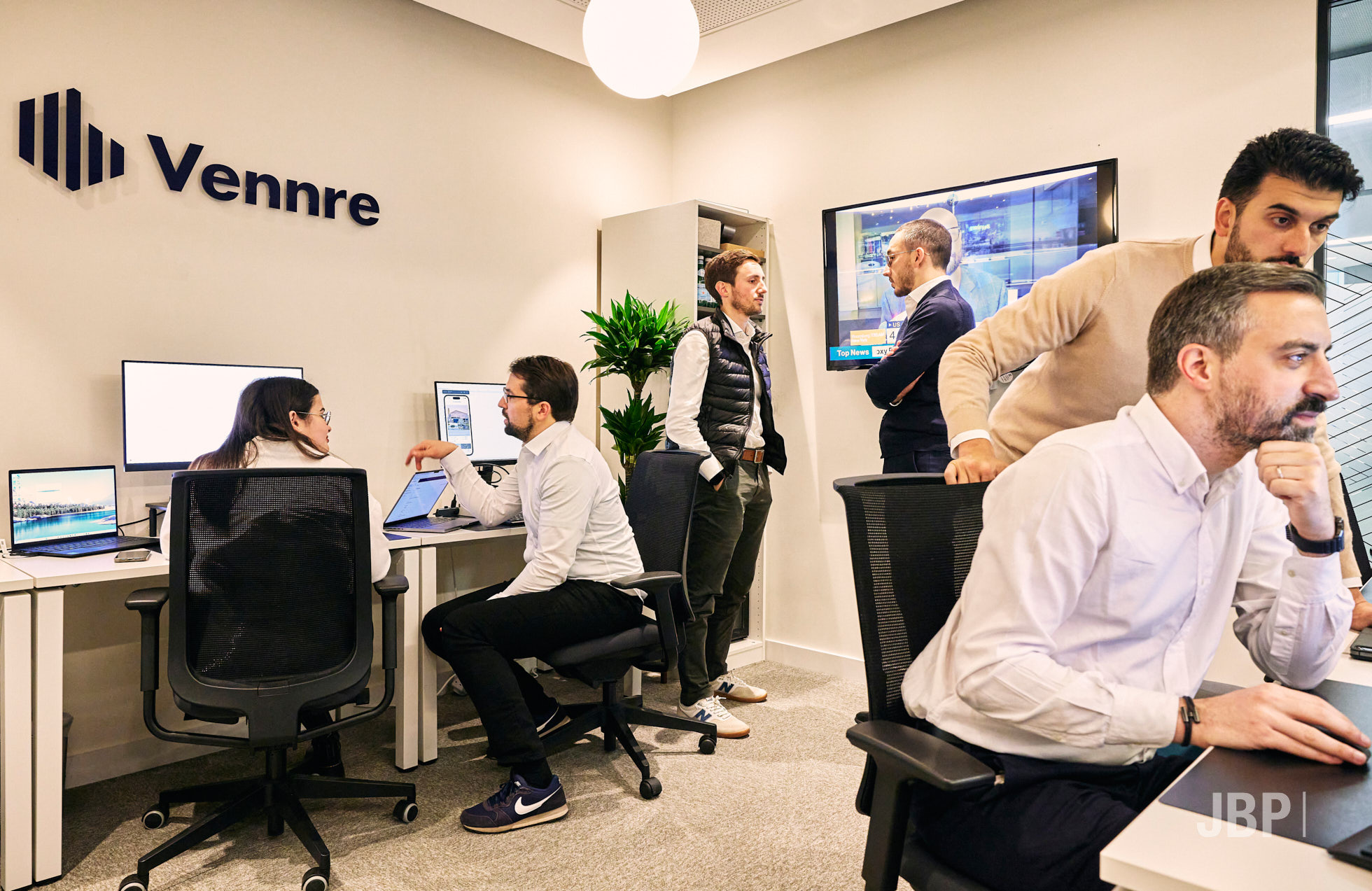 Corporate lifestyle photography showing a team of people in an office space all chatting, at computers with the company logo on the wall