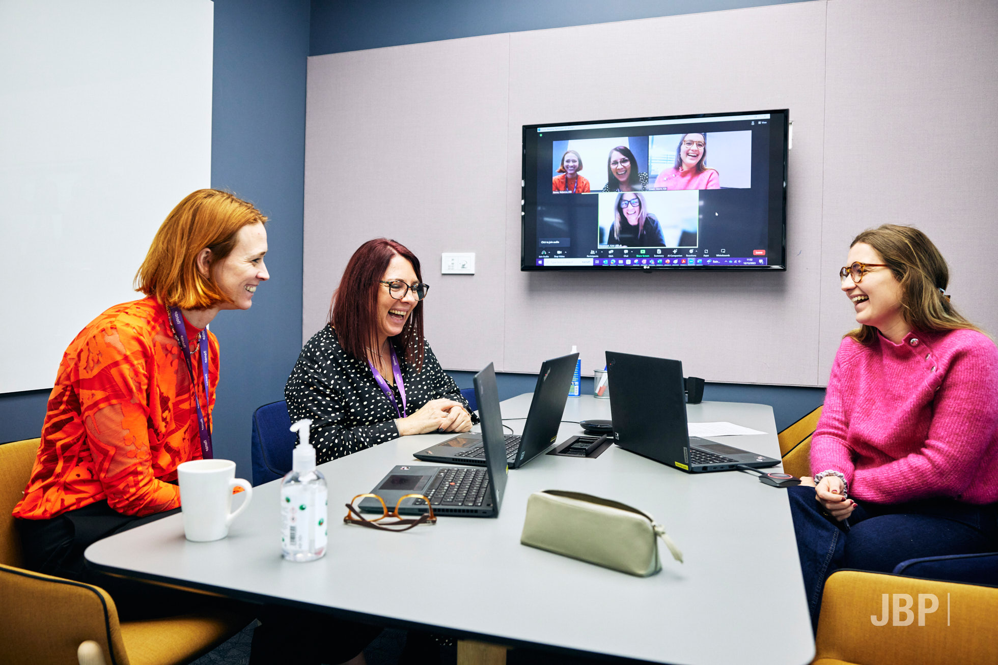 Corporate lifestyle photo of three women in a meeting room having an online meeting, all on laptops with their images on a tv screen