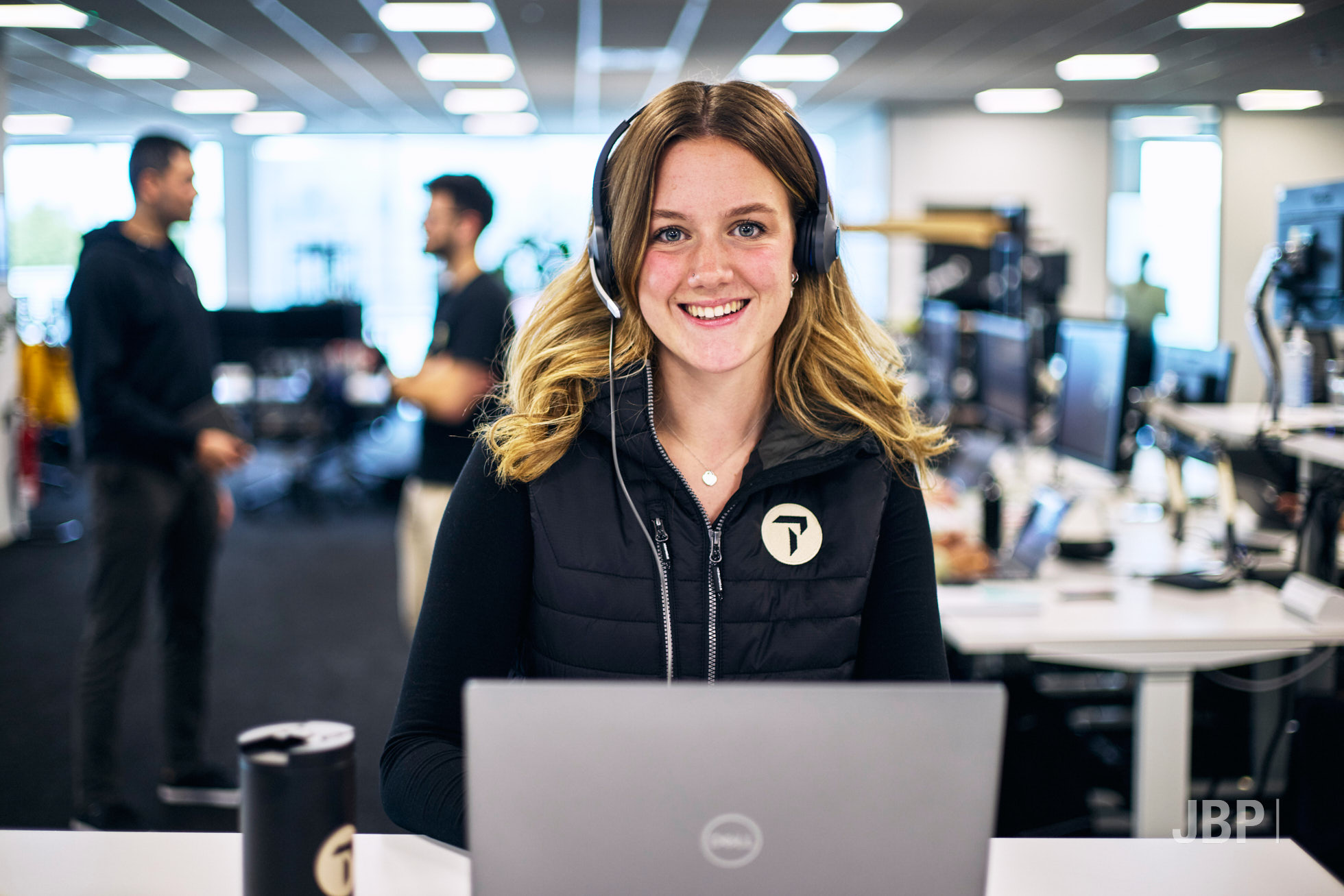 Women with a headset on at a laptop computer looking at the camera, with an office environment out of a focus in the background with two colleagues stood chatting.