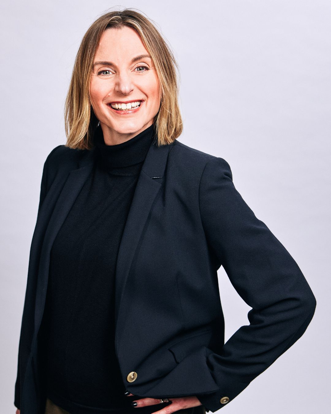 A smiling woman with shoulder-length blonde hair wearing a black turtleneck and black blazer, standing against a plain light-grey background with one hand on her hip.