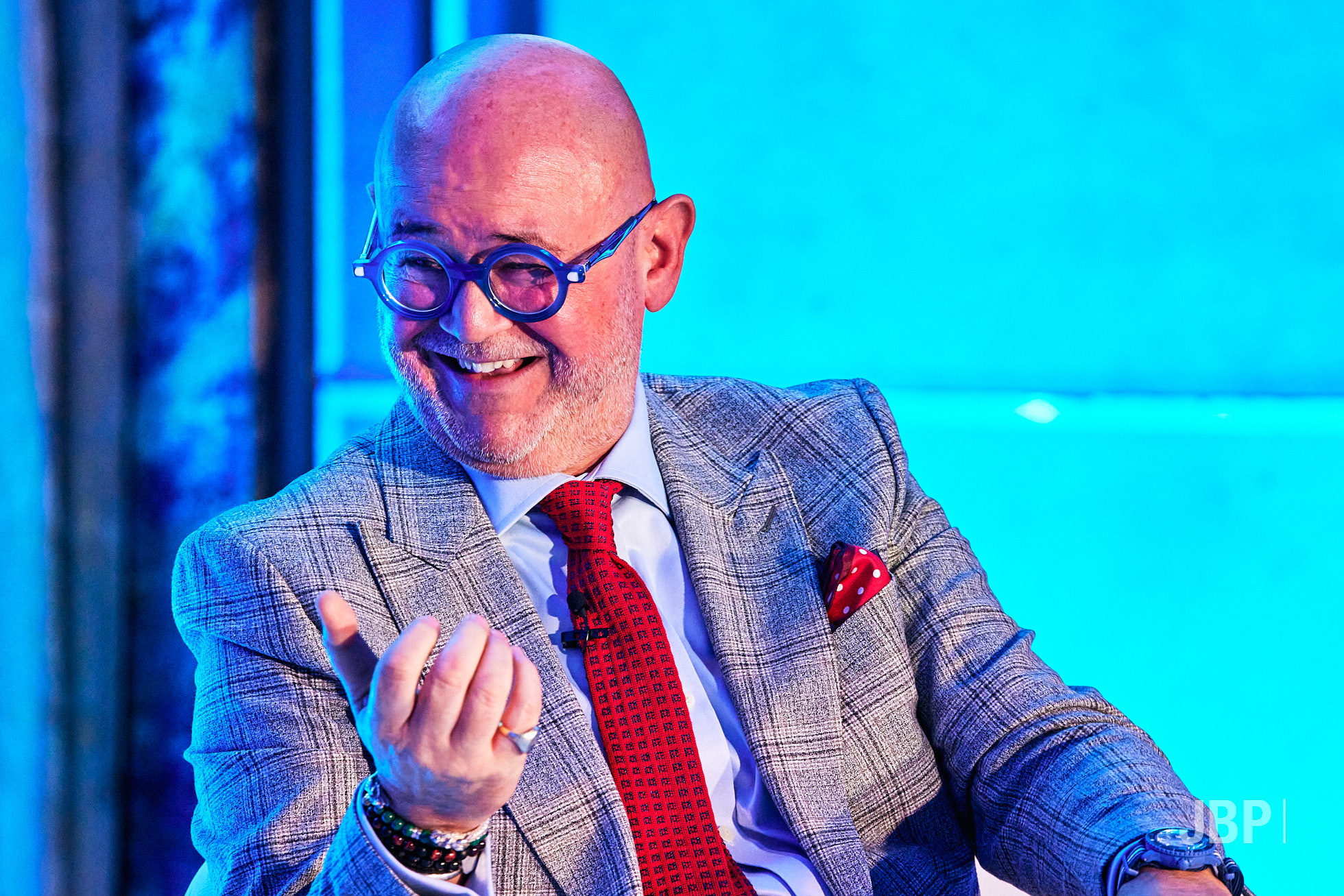 Event Photography by Jon Bradley A man with a grey beard wearing a gray suit, white shirt, and red tie sits in a blue-lit setting.