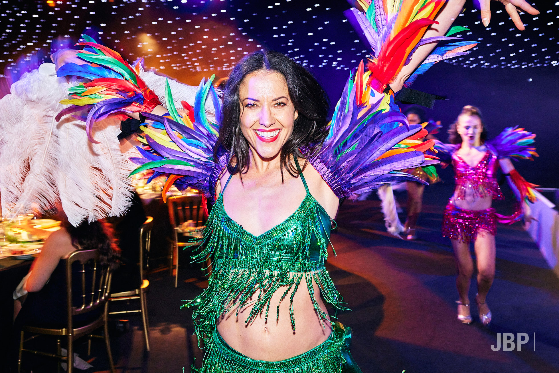 PR and event photography by Jon Bradley Photograohy Smiling woman in a green beaded fringe costume with multicolored feather wings, arms raised. Background dancers wear pink sequined outfits with feathers on a dark stage.