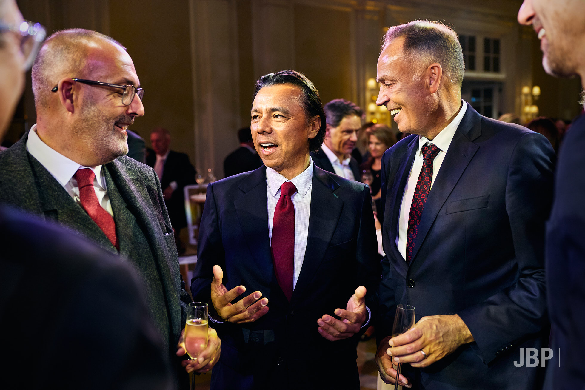 Event Photography by Jon Bradley Three men in suits chat and hold drinks at a formal indoor event, with other guests and ambient lighting in the background