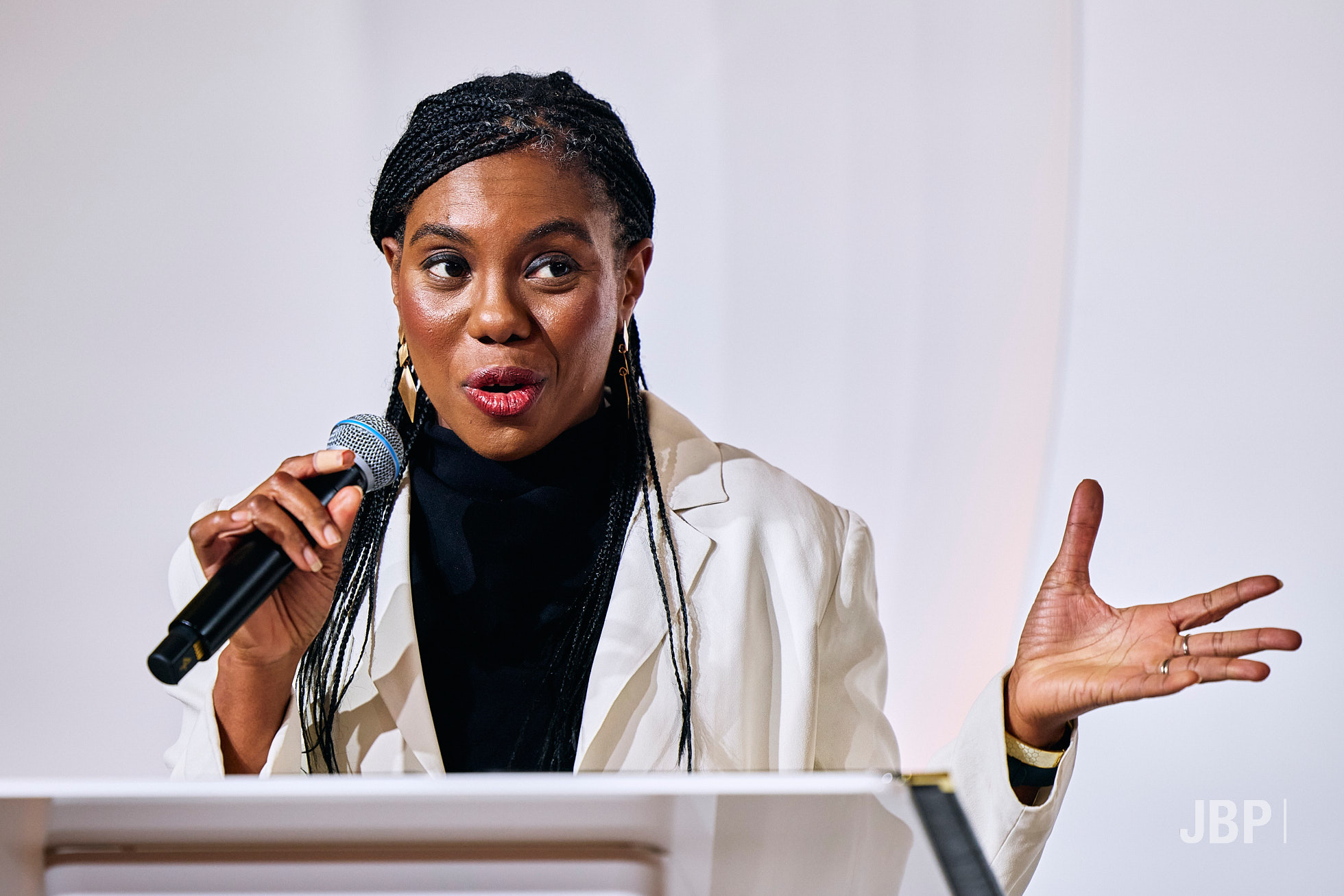 A woman with braided hair speaks animatedly into a microphone, wearing a white blazer and black top.