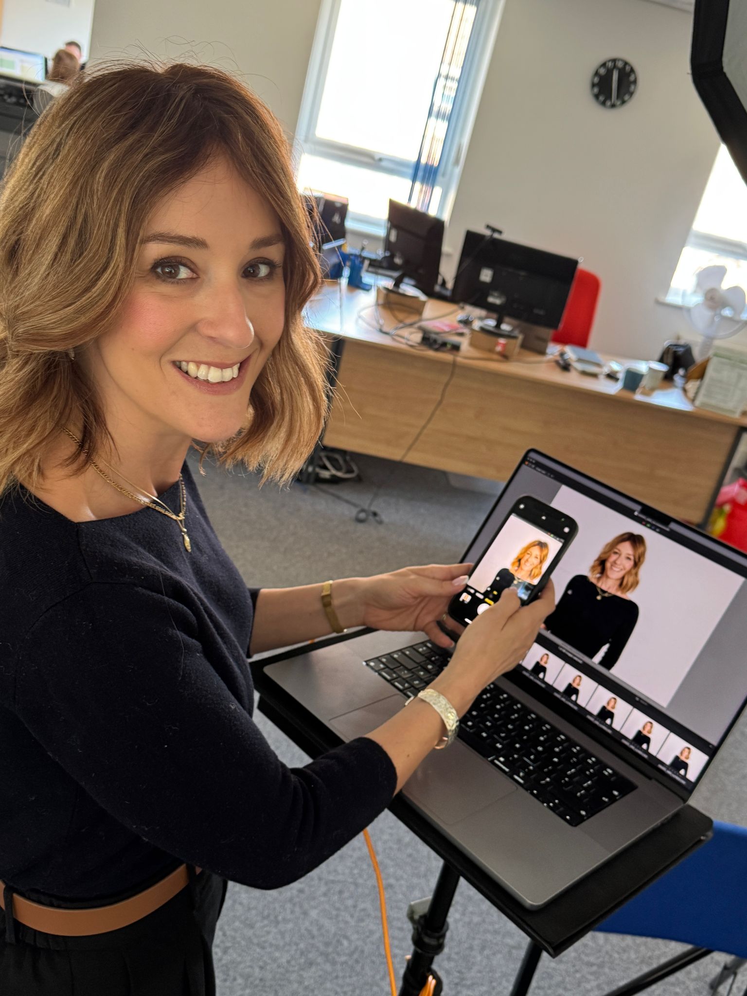 A smiling woman with shoulder-length hair in a dark top stands at a laptop on a stand in an office. She holds a phone displaying a portrait of herself.