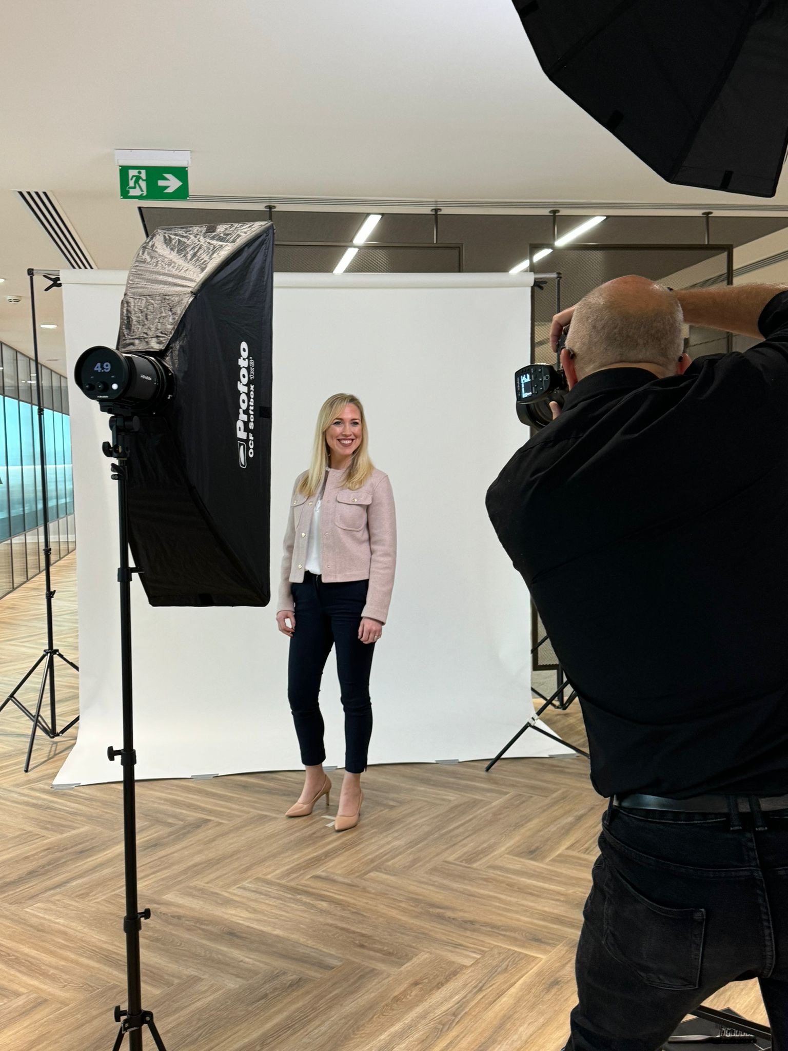 A blonde woman poses in a photography studio in front of a white backdrop, wearing a pale pink jacket, white top, dark jeans. A photographer uses large softbox lighting to shoot her, standing near the wooden floor.